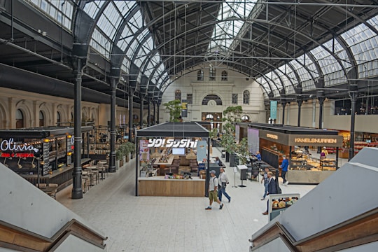 A spacious indoor marketplace with high, arched ceilings and large windows providing ample natural light. The scene includes various food stalls with visible signage, seating areas, and people walking or standing. The architecture has an industrial and historic style, combining metal beams and glass. Green plants add an element of nature to the space.