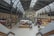 A spacious indoor marketplace with high, arched ceilings and large windows providing ample natural light. The scene includes various food stalls with visible signage, seating areas, and people walking or standing. The architecture has an industrial and historic style, combining metal beams and glass. Green plants add an element of nature to the space.
