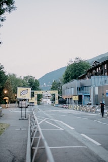 A quiet street scene in an urban area features signage and barriers suggesting preparation for a race or event, with buildings on one side and trees in the background. A finish line sign is visible in the distance, and a person is walking down the sidewalk.