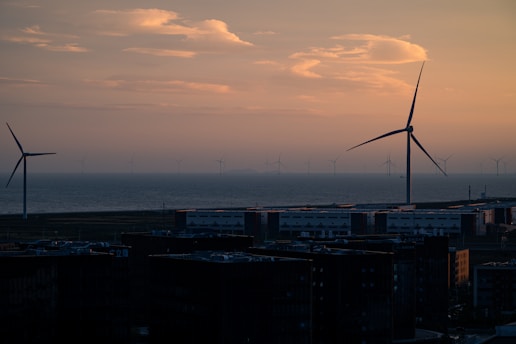 Wind turbines standing along the coastline during sunset, with a calm sea and a few scattered clouds overhead. Tall buildings are visible in the foreground, silhouetted against the colorful sky.