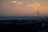 Wind turbines standing along the coastline during sunset, with a calm sea and a few scattered clouds overhead. Tall buildings are visible in the foreground, silhouetted against the colorful sky.