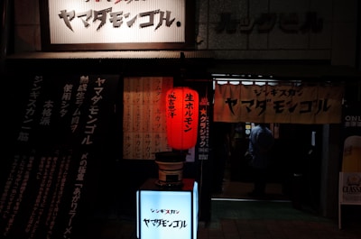 A dimly lit entrance to a restaurant adorned with Japanese signage, featuring a prominent red paper lantern and various banners with text. The doorway reveals a glimpse into the interior where a person stands.