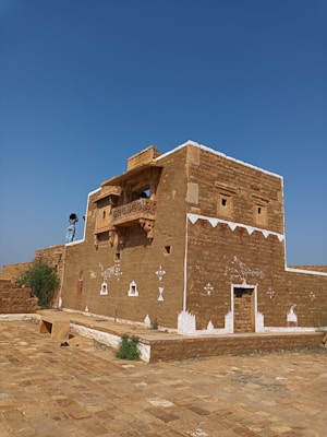 A traditional building constructed with earthy materials, featuring intricate white designs on the walls. The architecture includes small windows and a balcony with detailed carvings. The building is situated on a stone-paved surface under a clear blue sky, with some greenery visible around.