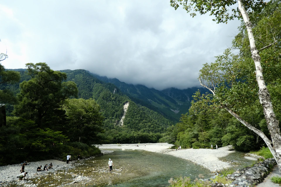 People stand near serene mountain landscape with flowing river surrounded by lush greenery outdoors