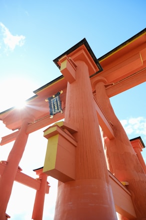 Bright orange wooden torii gate structure extends upwards towards a clear blue sky with some white clouds. The torii gate features traditional Japanese architectural elements and has a plaque with Japanese writing. Sunlight shines through the top left corner, creating a glowing effect.