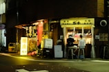 A small, cozy street-side restaurant with a glowing sign. Two people are seated at a counter, facing the open kitchen where food is being prepared. Warm lighting from the interior contrasts with the dark street outside.