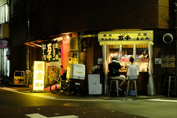 A small, cozy street-side restaurant with a glowing sign. Two people are seated at a counter, facing the open kitchen where food is being prepared. Warm lighting from the interior contrasts with the dark street outside.