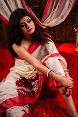 A woman dressed in a white and red traditional saree sits pensively, leaning on her knee. Her makeup is dramatic, featuring dark eyeshadow and a red bindi. The background features rich red and cream draped fabric, enhancing the warm and intense color scheme.