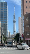 A tall broadcasting and observation tower stands prominently between modern buildings. The street below shows vehicles such as cars and trucks, along with a person on a bicycle. The sky is clear and blue, suggesting a sunny day.