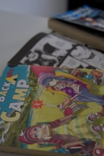 A stack of manga books featuring colorful anime-style illustrations on the covers. One book prominently displayed features two characters in a camping setting; a girl wearing a hat and scarf is eating a sandwich, and another character is sitting on a chair in a striped apron. The background shows an outdoor scene with a lake or river. In the upper right corner, there is a glimpse of another manga book with a darker cover and text in Japanese, hinting at black and white pages inside.