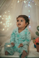 A young child wearing light blue pajamas sits in a cozy setting adorned with soft, ambient fairy lights. The child holds a small toy and appears to be looking slightly upward with a curious and engaging expression. The background includes a sheer curtain and a plush toy shaped like a sea creature, adding to the whimsical atmosphere.