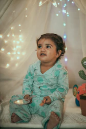 A young child wearing light blue pajamas sits in a cozy setting adorned with soft, ambient fairy lights. The child holds a small toy and appears to be looking slightly upward with a curious and engaging expression. The background includes a sheer curtain and a plush toy shaped like a sea creature, adding to the whimsical atmosphere.