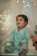 A young child wearing light blue pajamas sits in a cozy setting adorned with soft, ambient fairy lights. The child holds a small toy and appears to be looking slightly upward with a curious and engaging expression. The background includes a sheer curtain and a plush toy shaped like a sea creature, adding to the whimsical atmosphere.