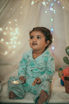 A young child wearing light blue pajamas sits in a cozy setting adorned with soft, ambient fairy lights. The child holds a small toy and appears to be looking slightly upward with a curious and engaging expression. The background includes a sheer curtain and a plush toy shaped like a sea creature, adding to the whimsical atmosphere.