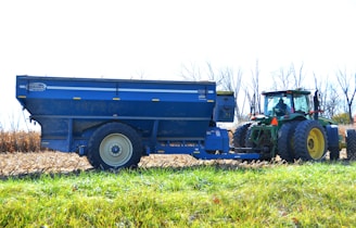 A tractor is attached to a large blue agricultural trailer in a field. The tractor has large wheels and is mostly green with yellow rims. The field appears to have been recently harvested, with dried cornstalks visible. Bare trees line the background, and the sky is clear and bright.