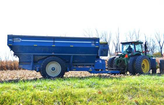 A tractor is attached to a large blue agricultural trailer in a field. The tractor has large wheels and is mostly green with yellow rims. The field appears to have been recently harvested, with dried cornstalks visible. Bare trees line the background, and the sky is clear and bright.