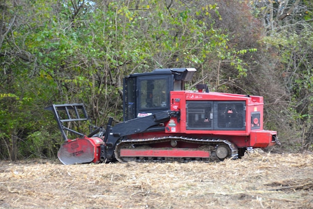 A red tracked vehicle, possibly a forestry mulcher, is situated in a natural setting surrounded by dense green foliage and dried grass. The vehicle has a sturdy build with a large cutting attachment at the front, designed for clearing vegetation.