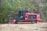 A red tracked vehicle, possibly a forestry mulcher, is situated in a natural setting surrounded by dense green foliage and dried grass. The vehicle has a sturdy build with a large cutting attachment at the front, designed for clearing vegetation.