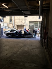 A group of people gathered in an alleyway or garage space with two cars parked. The car in the foreground is a black, four-door sedan with its doors open, and people are either standing around or sitting on it. The background features a brick building facade with large windows and some industrial elements like ventilation ducts and utility fixtures.