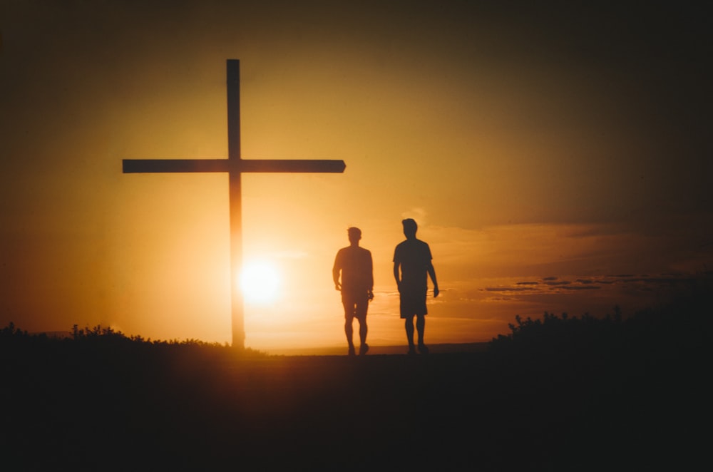 two people standing in front of a cross at sunset
