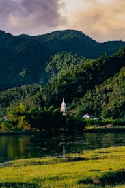 A serene landscape featuring a tranquil body of water in the foreground, bordered by lush green grass. In the background, a church with a tall white steeple nestles among dense green foliage, set against a backdrop of forested hills. The sky is partly cloudy, with soft, warm hues suggesting a setting or rising sun.