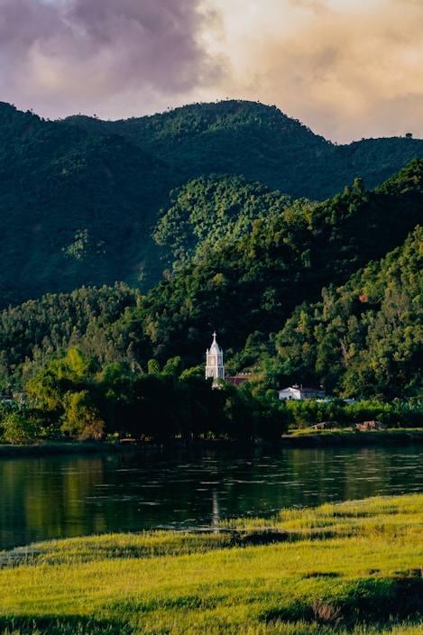 A serene landscape featuring a tranquil body of water in the foreground, bordered by lush green grass. In the background, a church with a tall white steeple nestles among dense green foliage, set against a backdrop of forested hills. The sky is partly cloudy, with soft, warm hues suggesting a setting or rising sun.