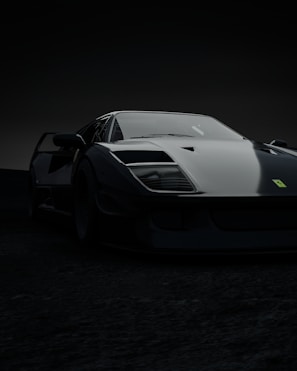 A sleek black sports car is captured in low light, highlighting its aerodynamic shape and glossy surface. The car's front bumper and headlights are prominently visible, reflecting a faint light source. The background is dark and blurry, focusing attention on the car.