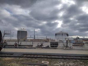 Industrial site with large cylindrical storage tanks and surrounding infrastructure. The sky is overcast with thick gray clouds, creating a moody atmosphere. There are multiple pipes, rail tracks, and industrial equipment present.