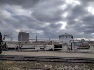 Industrial site with large cylindrical storage tanks and surrounding infrastructure. The sky is overcast with thick gray clouds, creating a moody atmosphere. There are multiple pipes, rail tracks, and industrial equipment present.