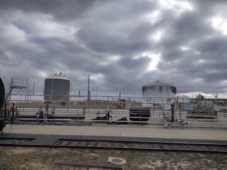 Industrial site with large cylindrical storage tanks and surrounding infrastructure. The sky is overcast with thick gray clouds, creating a moody atmosphere. There are multiple pipes, rail tracks, and industrial equipment present.