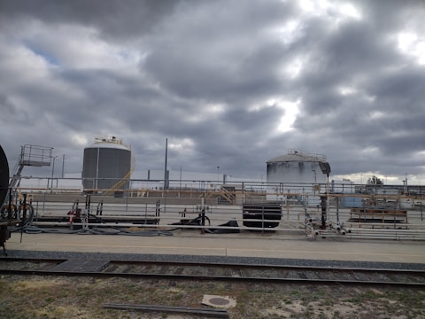 Industrial site with large cylindrical storage tanks and surrounding infrastructure. The sky is overcast with thick gray clouds, creating a moody atmosphere. There are multiple pipes, rail tracks, and industrial equipment present.