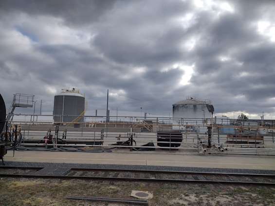 Industrial site with large cylindrical storage tanks and surrounding infrastructure. The sky is overcast with thick gray clouds, creating a moody atmosphere. There are multiple pipes, rail tracks, and industrial equipment present.