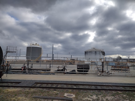 Industrial site with large cylindrical storage tanks and surrounding infrastructure. The sky is overcast with thick gray clouds, creating a moody atmosphere. There are multiple pipes, rail tracks, and industrial equipment present.