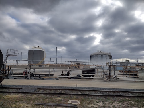 Industrial site with large cylindrical storage tanks and surrounding infrastructure. The sky is overcast with thick gray clouds, creating a moody atmosphere. There are multiple pipes, rail tracks, and industrial equipment present.