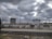 Industrial site with large cylindrical storage tanks and surrounding infrastructure. The sky is overcast with thick gray clouds, creating a moody atmosphere. There are multiple pipes, rail tracks, and industrial equipment present.