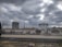 Industrial site with large cylindrical storage tanks and surrounding infrastructure. The sky is overcast with thick gray clouds, creating a moody atmosphere. There are multiple pipes, rail tracks, and industrial equipment present.