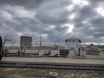 Industrial site with large cylindrical storage tanks and surrounding infrastructure. The sky is overcast with thick gray clouds, creating a moody atmosphere. There are multiple pipes, rail tracks, and industrial equipment present.