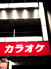 Brightly lit karaoke sign with bold white Japanese characters on a vibrant red background, contrasted with the dimly lit building facade behind it.