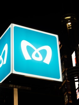 A large, illuminated outdoor sign featuring a stylized logo resembling the letter 'M' is prominently displayed against a dark night sky. The sign is bright and stands out due to its blue background and white logo. The setting appears to be urban, with parts of a building and additional lighting visible.