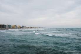 A coastal cityscape with numerous surfers in action on the waves of a vast, choppy ocean. The shoreline is lined with mid-rise buildings, suggesting a developed urban area. The sky is overcast, contributing to a somewhat subdued atmosphere.