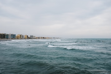 A coastal cityscape with numerous surfers in action on the waves of a vast, choppy ocean. The shoreline is lined with mid-rise buildings, suggesting a developed urban area. The sky is overcast, contributing to a somewhat subdued atmosphere.
