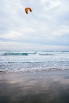 A kite surfer navigates the ocean waves with an orange and black kite soaring high above. The sea is choppy with visible waves while the sky is overcast, casting a muted light over the scene. The sandy beach in the foreground is wet, reflecting the grayish tones of the sky.
