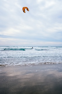 A kite surfer navigates the ocean waves with an orange and black kite soaring high above. The sea is choppy with visible waves while the sky is overcast, casting a muted light over the scene. The sandy beach in the foreground is wet, reflecting the grayish tones of the sky.