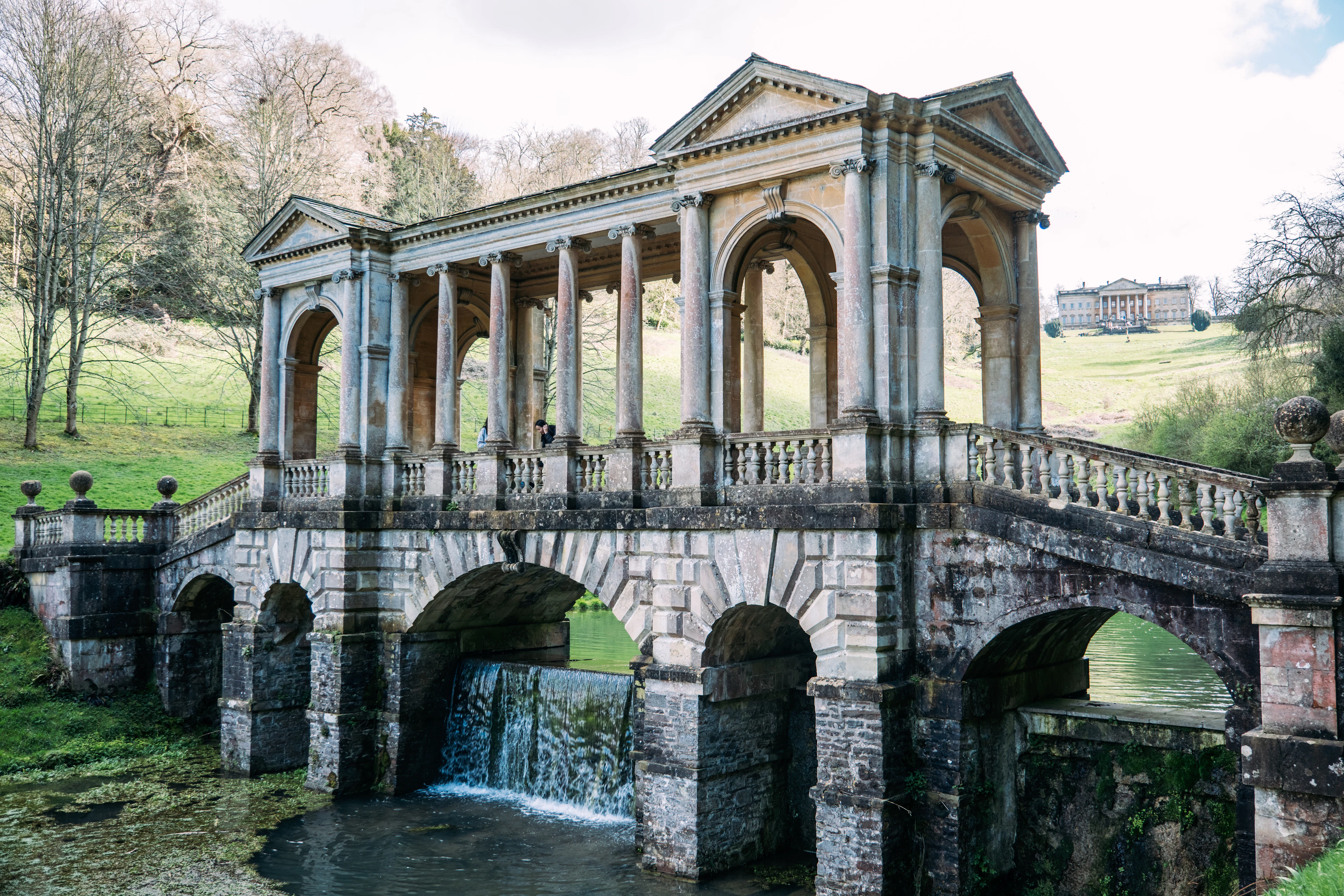 The Palladian Bridge at Prior Park in Bath, its colonnade and pediments standing above a weir spilling into the lake, with the mansion on the green hillside beyond.