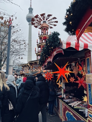A bustling outdoor market scene featuring vibrant holiday decorations, including illuminated star-shaped ornaments hanging from stalls adorned with festive greenery. A crowd of people, dressed in winter clothing, explores the market, with a towering structure and a prominent television tower in the background.
