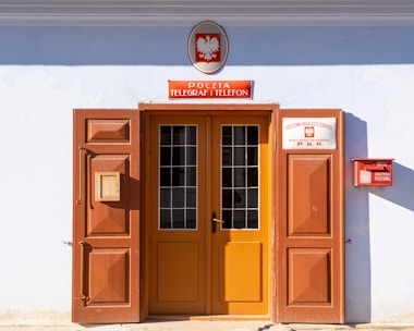 A doorway with large wooden doors, one of which is open, revealing a glass-panelled door behind it. Above the entrance, there is a sign with text in a foreign language and an emblem featuring an eagle. A red mailbox is attached to the right wall, along with another sign detailing postal savings.