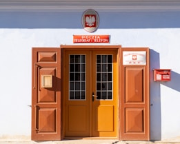 A doorway with large wooden doors, one of which is open, revealing a glass-panelled door behind it. Above the entrance, there is a sign with text in a foreign language and an emblem featuring an eagle. A red mailbox is attached to the right wall, along with another sign detailing postal savings.