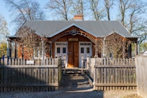 A charming wooden house with a gabled roof is surrounded by a wooden picket fence. Leafless trees stand in front, and a pathway leads to the entrance. The sign above the door reads 'RESTALACIA,' indicating a restaurant or similar establishment.