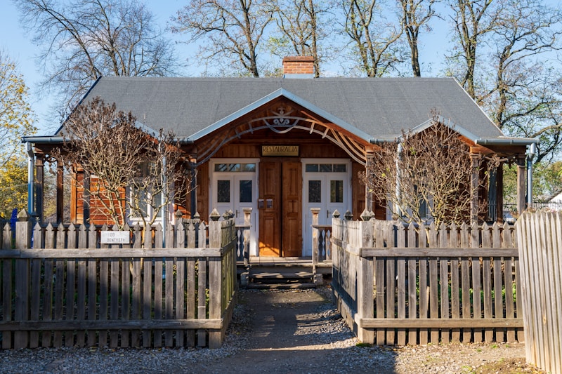 A charming wooden house with a gabled roof is surrounded by a wooden picket fence. Leafless trees stand in front, and a pathway leads to the entrance. The sign above the door reads 