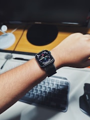 A person wearing a smartwatch on their wrist, with a digital display showing the time and various widgets. In the background, there is a wooden desk with a computer monitor, a round speaker, and a keyboard partially visible.